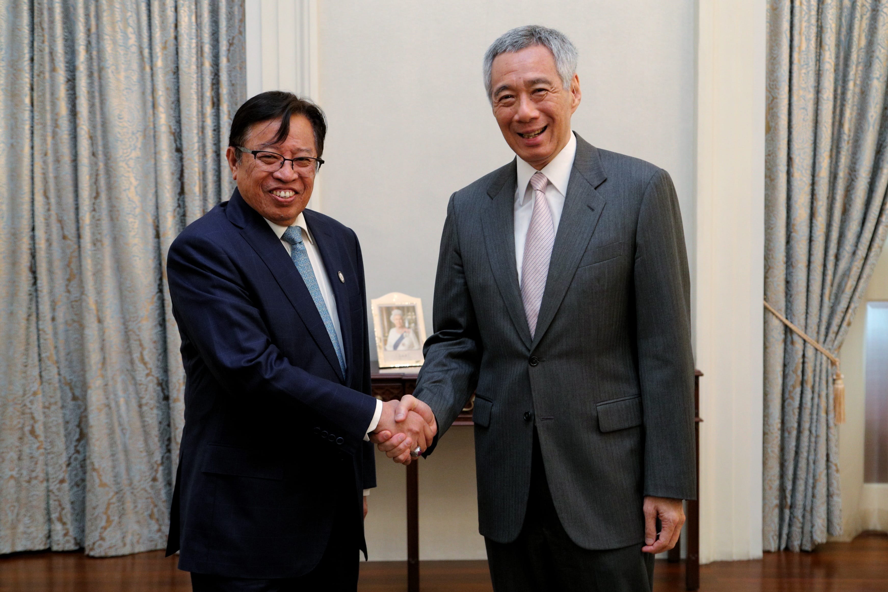 Two men in suits shake hands in a room with curtains and a framed photo.